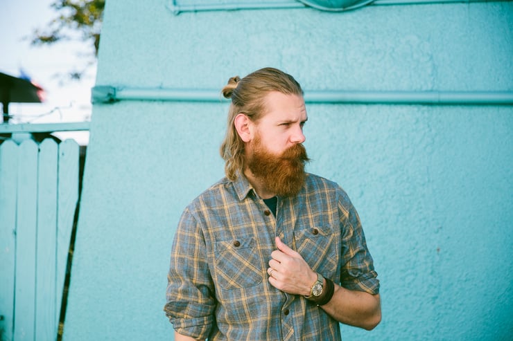 A gentleman with a robust red, beard and plaid shirt poses in front of a blue wall.